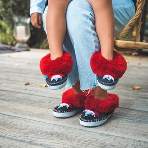 May include: A pair of black moccasin-style slippers with red fur trim. The slippers have a white sole and a decorative design in white and red. The image shows the slippers being worn by a person.