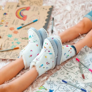 May include: A pair of white fuzzy boots with colorful drawings on them. The boots are worn by a child who is sitting on a rug with several colorful markers.
