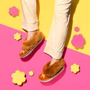 Peut inclure: Une paire de pantoufles en fourrure brune avec une semelle en daim. Les pantoufles sont portées par une personne vêtue d'un pantalon beige sur un fond jaune et rose.