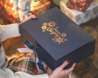 May include: A navy blue gift box with gold lettering that says "Merry Christmas" and decorative snowflakes. The box is tied with a blue and pink ribbon. The box is held in front of a Christmas tree.