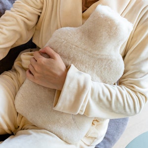 May include: A white faux fur hot water bottle being held by a person wearing a cream coloured robe.
