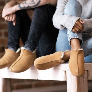 May include: Two pairs of tan suede slippers with a white sheepskin lining. The slippers are worn by two people sitting on a wooden bench.