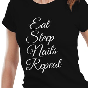 May include: Black t-shirt with the white text "Eat Sleep Nails Repeat" in a cursive font. The shirt is a crew neck style and is being worn.