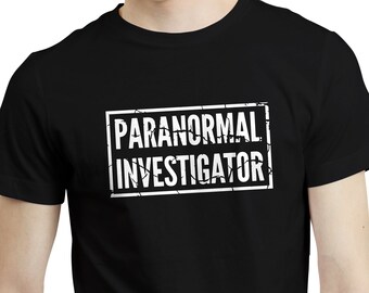 Może przedstawiać: Czarny t-shirt z napisem "PARANORMAL INVESTIGATOR" w białym, postarzanym kroju wewnątrz białego prostokąta. Koszulka z krótkim rękawem wykonana jest z miękkiego materiału.