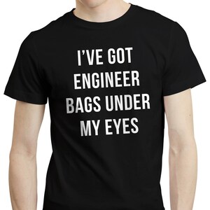 Può includere: T-shirt nero con la scritta bianca "I'VE GOT ENGINEER BAGS UNDER MY EYES". La maglietta è a girocollo e sembra essere realizzata in un materiale morbido. Il testo è centrato sulla parte anteriore della maglietta.