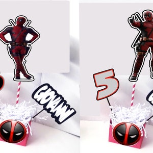 May include: Two pink boxes with a Deadpool character design. Each box has a white paper shred filler.  One box has a black number 5 and a black and white 'GIOVANNI' sign. The other box has a gray number 5 and a black and white 'GIOVANNI' sign. Both boxes have a Deadpool character on a stick.