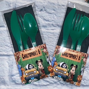 May include: Two sets of green plastic cutlery, a knife, fork, and spoon, are packaged in clear plastic bags with black napkins. Each bag has a colorful label with a cartoon image of Mickey Mouse in a safari outfit driving a jeep with jungle animals. The label also says "Christopher's 6".