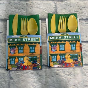 May include: Two sets of yellow plastic cutlery, including a knife, fork, and spoon, are presented on green napkins. Each set is packaged with a card featuring the "MEKHI STREET" logo and Sesame Street characters. The background is a gray brick wall.