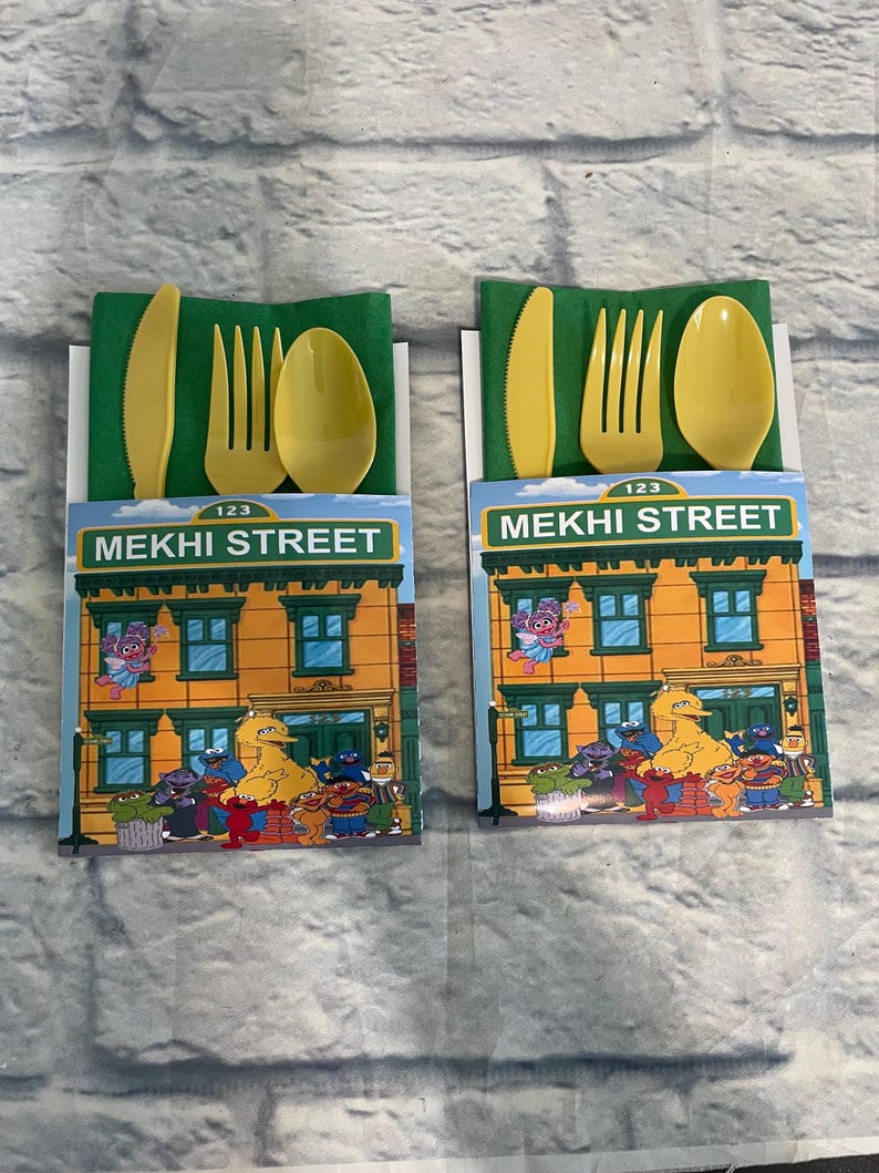 May include: Two sets of yellow plastic cutlery, including a knife, fork, and spoon, are presented on green napkins. Each set is packaged with a card featuring the "MEKHI STREET" logo and Sesame Street characters. The background is a gray brick wall.