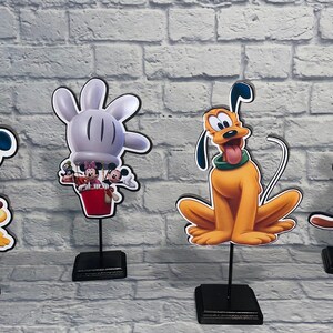 May include: Four Disney character themed centerpieces featuring Mickey Mouse, Donald Duck, Goofy, and Pluto. Each character is printed on a cardboard cutout and mounted on a black stand.
