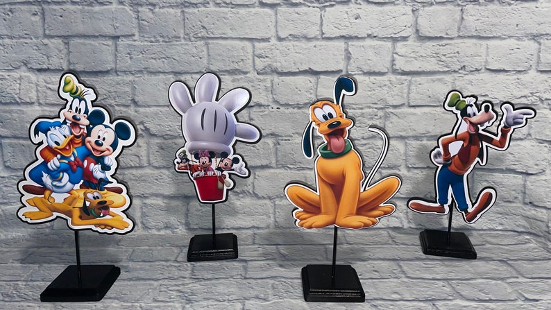 May include: Four Disney character themed centerpieces featuring Mickey Mouse, Donald Duck, Goofy, and Pluto. Each character is printed on a cardboard cutout and mounted on a black stand.