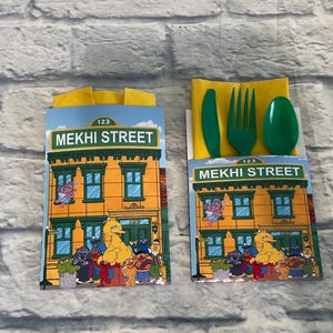 May include: Two party favor boxes featuring a Sesame Street theme. Each box displays a building facade with the text "MEKHI STREET" and various Sesame Street characters. One box contains a green plastic knife, fork, and spoon.
