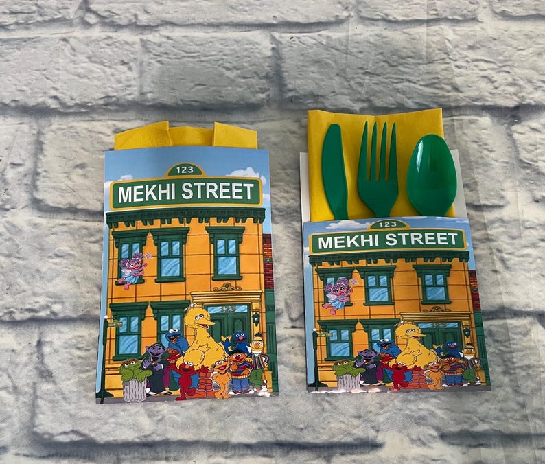May include: Two party favor boxes featuring a Sesame Street theme. Each box displays a building facade with the text "MEKHI STREET" and various Sesame Street characters. One box contains a green plastic knife, fork, and spoon.