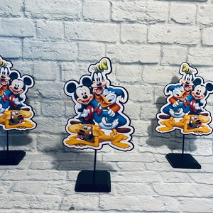 May include: Three tabletop centerpieces featuring Mickey Mouse, Donald Duck, Goofy, and Pluto. The characters are depicted in a cartoon style with bright colors and playful expressions. Each centerpiece is mounted on a black stand.