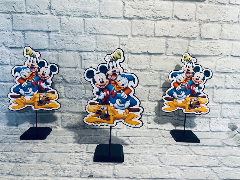 May include: Three tabletop centerpieces featuring Mickey Mouse, Donald Duck, Goofy, and Pluto. The characters are depicted in a cartoon style with bright colors and playful expressions. Each centerpiece is mounted on a black stand.