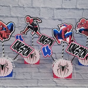 以下が含まれることがあります： 誕生日パーティー用のスパイダーマンをテーマにしたセンターピースが5つ。各センターピースには、クモの巣のデザインが施された赤と青の段ボール箱、黒と赤のスパイダーマンのフィギュア、黒と白の数字4、そして「Kingston 4」と書かれた黒と白の看板が付いています。
