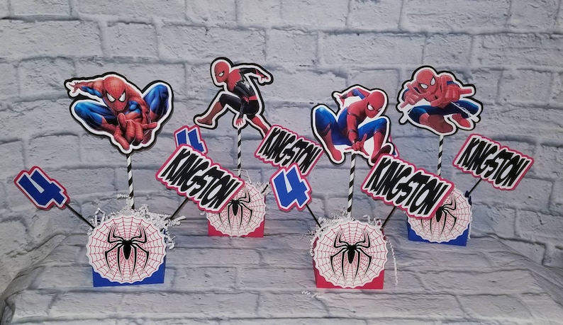 以下が含まれることがあります： 誕生日パーティー用のスパイダーマンをテーマにしたセンターピースが5つ。各センターピースには、クモの巣のデザインが施された赤と青の段ボール箱、黒と赤のスパイダーマンのフィギュア、黒と白の数字4、そして「Kingston 4」と書かれた黒と白の看板が付いています。
