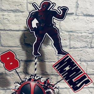 May include: A red and black Deadpool themed centerpiece with a red cube featuring a Deadpool mask, a black and white striped straw, a red number 8, and a black sign with the name "Nicholas" in white letters.