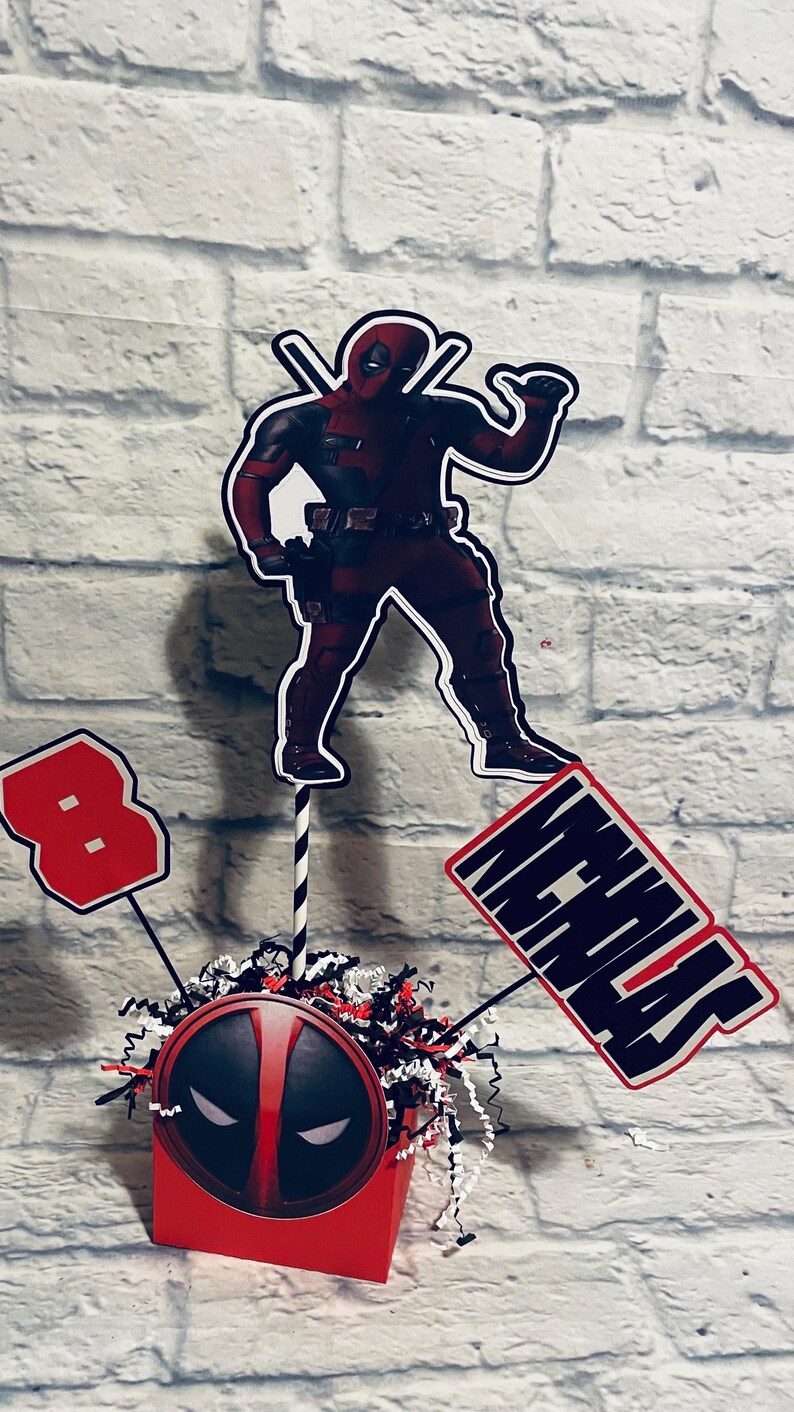 May include: A red and black Deadpool themed centerpiece with a red cube featuring a Deadpool mask, a black and white striped straw, a red number 8, and a black sign with the name "Nicholas" in white letters.