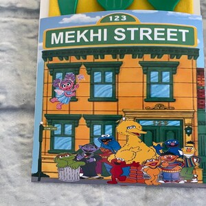 May include: A colorful illustration of a building with the text "MEKHI STREET" and the number "123". The image features various cartoon characters, including Big Bird, Elmo, and Cookie Monster. The building is orange with green window frames.