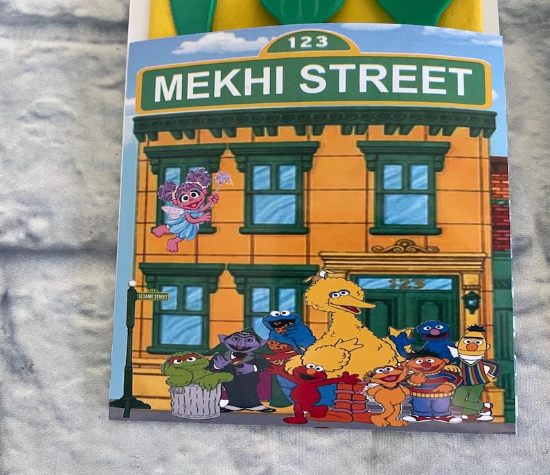 May include: A colorful illustration of a building with the text "MEKHI STREET" and the number "123". The image features various cartoon characters, including Big Bird, Elmo, and Cookie Monster. The building is orange with green window frames.