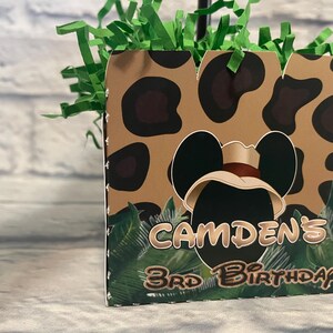 Puede incluir: Una caja de regalo cuadrada de cart&oacute;n con un patr&oacute;n de leopardo en color canela y negro, gr&aacute;ficos de hojas de palma verdes y una silueta central negra de orejas de rat&oacute;n con un sombrero de safari. El texto dice CAMDEN'S 3RD BIRTHDAY en marr&oacute;n.