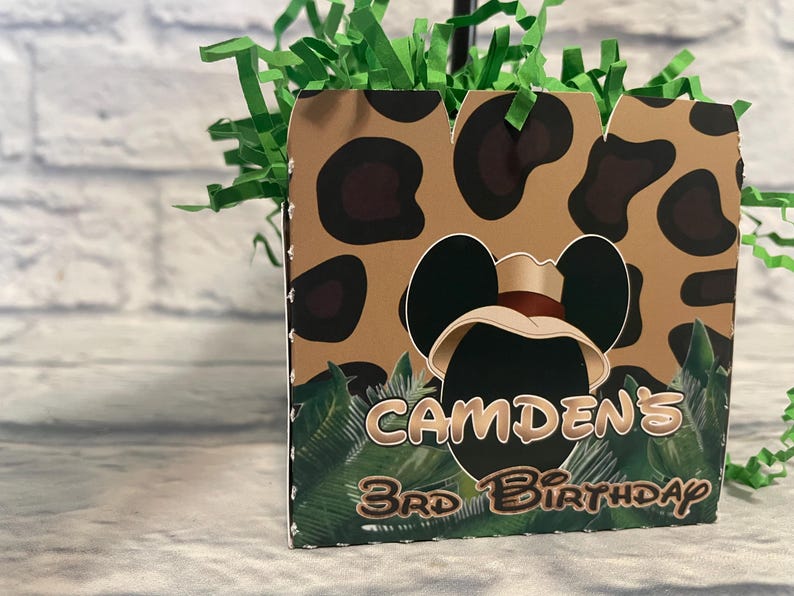 Puede incluir: Una caja de regalo cuadrada de cart&oacute;n con un patr&oacute;n de leopardo en color canela y negro, gr&aacute;ficos de hojas de palma verdes y una silueta central negra de orejas de rat&oacute;n con un sombrero de safari. El texto dice CAMDEN'S 3RD BIRTHDAY en marr&oacute;n.