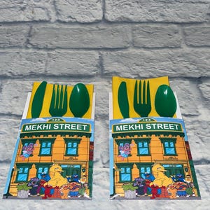 May include: Two sets of children's cutlery, each with a green knife, fork, and spoon, atop a yellow napkin. The packaging features a colorful illustration of a building with the text "MEKHI STREET" and Sesame Street characters.
