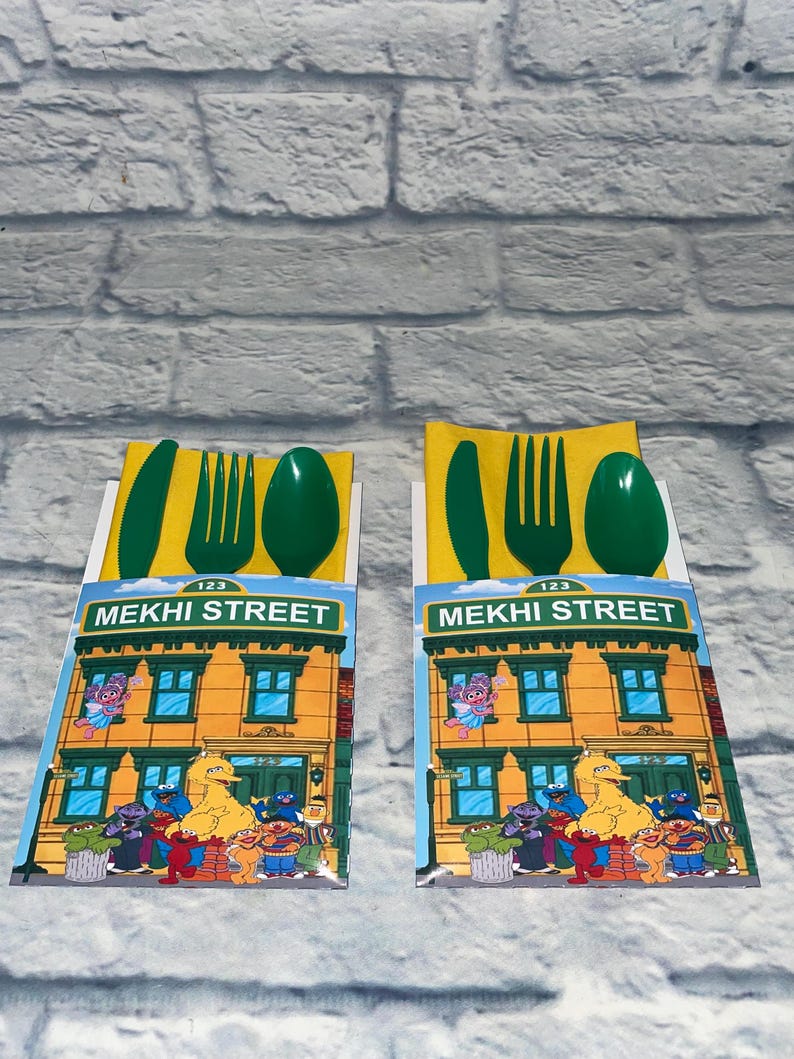 May include: Two sets of children's cutlery, each with a green knife, fork, and spoon, atop a yellow napkin. The packaging features a colorful illustration of a building with the text "MEKHI STREET" and Sesame Street characters.