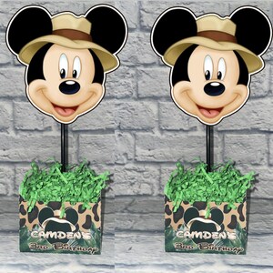 Puede incluir: Dos centros de mesa con tem&aacute;tica de safari de Mickey Mouse con un recorte de Mickey usando un sombrero de explorador color canela en un palo negro. La base es una caja cuadrada con estampado de leopardo, pasto de papel verde y texto Camden's 3rd Birthday.