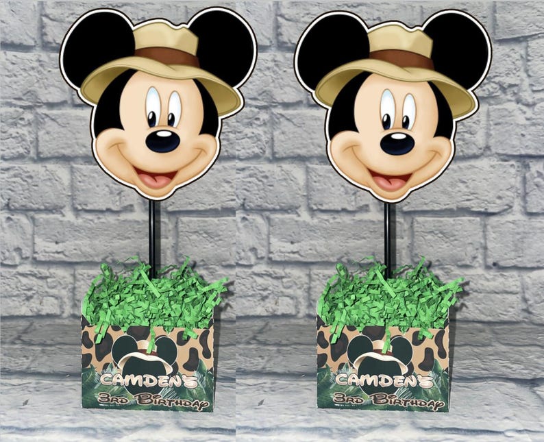 Puede incluir: Dos centros de mesa con tem&aacute;tica de safari de Mickey Mouse con un recorte de Mickey usando un sombrero de explorador color canela en un palo negro. La base es una caja cuadrada con estampado de leopardo, pasto de papel verde y texto Camden's 3rd Birthday.