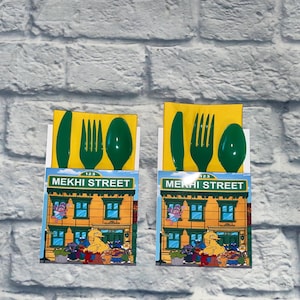 May include: Two Sesame Street-themed party favor sets. Each set includes a green plastic knife, fork, and spoon on a yellow background. The sets feature a colorful illustration of the Sesame Street building with the text "MEKHI STREET".