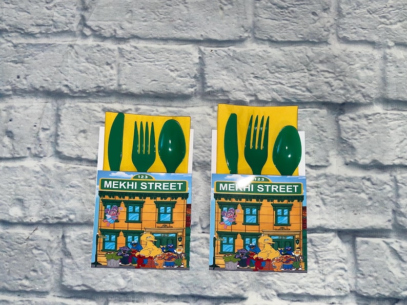 May include: Two Sesame Street-themed party favor sets. Each set includes a green plastic knife, fork, and spoon on a yellow background. The sets feature a colorful illustration of the Sesame Street building with the text "MEKHI STREET".