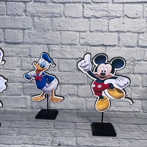 May include: Four cardboard cutouts of Disney characters on black stands. The characters are Daisy Duck in a purple dress, Donald Duck in a blue sailor shirt, Mickey Mouse in red shorts, and Minnie Mouse in a red polka dot dress.