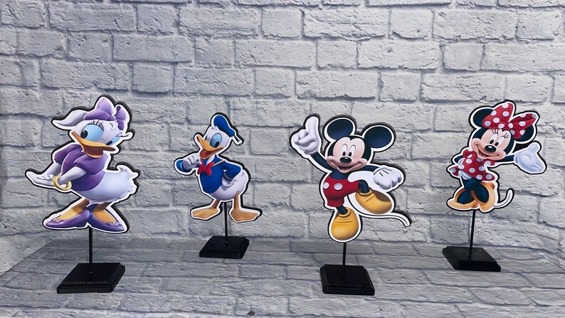 May include: Four cardboard cutouts of Disney characters on black stands. The characters are Daisy Duck in a purple dress, Donald Duck in a blue sailor shirt, Mickey Mouse in red shorts, and Minnie Mouse in a red polka dot dress.