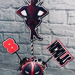 May include: A red and black Deadpool themed centerpiece with a red number 8, a black and white Deadpool mask, and a sign that says "Nicholas".