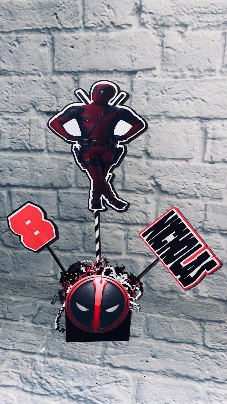May include: A red and black Deadpool themed centerpiece with a red number 8, a black and white Deadpool mask, and a sign that says "Nicholas".