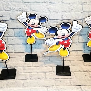 May include: Four black tabletop stands with Mickey Mouse cutouts. Each cutout features Mickey Mouse with a red shirt, black shorts, and yellow shoes. The cutouts are on black stands.