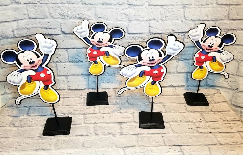 May include: Four black tabletop stands with Mickey Mouse cutouts. Each cutout features Mickey Mouse with a red shirt, black shorts, and yellow shoes. The cutouts are on black stands.