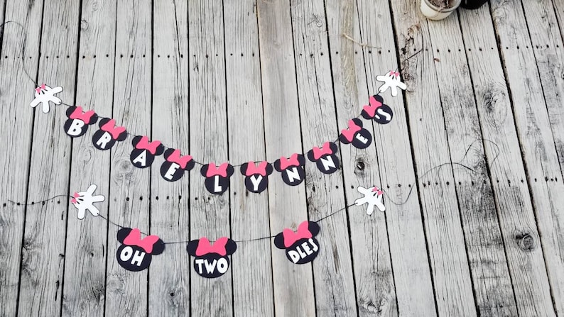 May include: A black and pink Minnie Mouse themed banner with the text "Braelynn is two" and white Mickey Mouse hands on each end.
