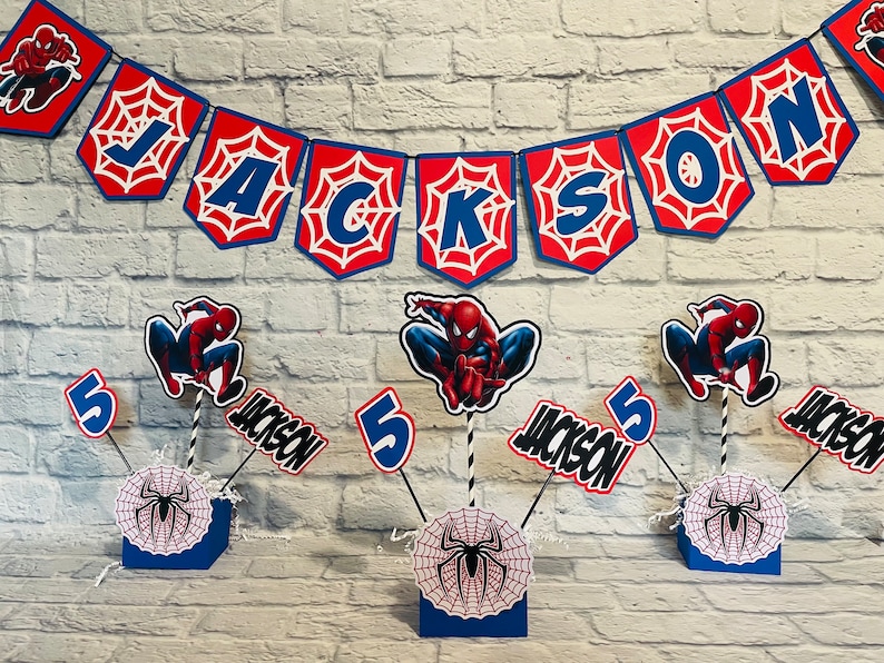 以下が含まれることがあります： スパイダーマンをテーマにしたパーティーの飾り付け。赤と青の旗に「JACKSON」と書いてあり、蜘蛛の巣のデザイン、赤と青のスパイダーマンのフィギュア、数字の「5」、そして「JACKSON」という名前が書かれたセンターピースが3つあります。