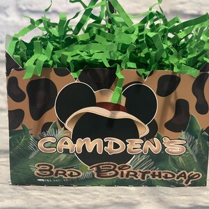 Puede incluir: Una caja rectangular de cart&oacute;n llena de papel triturado verde. El exterior presenta un estampado animal marr&oacute;n y negro, una silueta de orejas de rat&oacute;n negras con un sombrero beige y el texto CAMDEN'S 3RD BIRTHDAY en una fuente estilizada.
