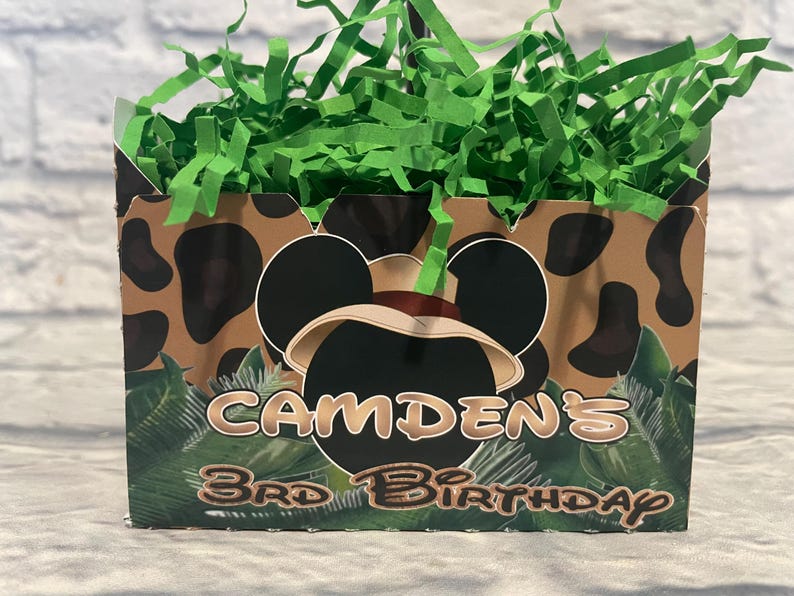 Puede incluir: Una caja rectangular de cart&oacute;n llena de papel triturado verde. El exterior presenta un estampado animal marr&oacute;n y negro, una silueta de orejas de rat&oacute;n negras con un sombrero beige y el texto CAMDEN'S 3RD BIRTHDAY en una fuente estilizada.