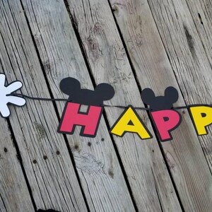 May include: A black, red, and yellow banner with the word "HAPPY" spelled out in large letters. Each letter has a Mickey Mouse head on top. A white Mickey Mouse glove is attached to the left side of the banner.