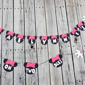 May include: A black and pink Minnie Mouse themed banner with the text "Braelynn is Two" and white Mickey Mouse hands on either end.