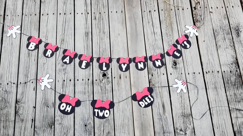 May include: A black and pink Minnie Mouse themed banner with the text "Braelynn is Two" and white Mickey Mouse hands on either end.
