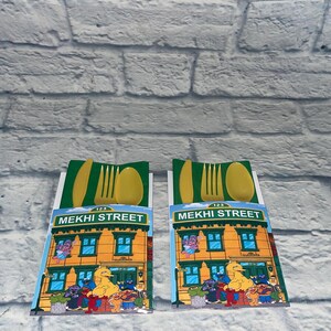 May include: Two sets of yellow plastic cutlery, each packaged with a green and white card featuring a Sesame Street building illustration. The card reads "MEKHI STREET" and includes various Sesame Street characters.