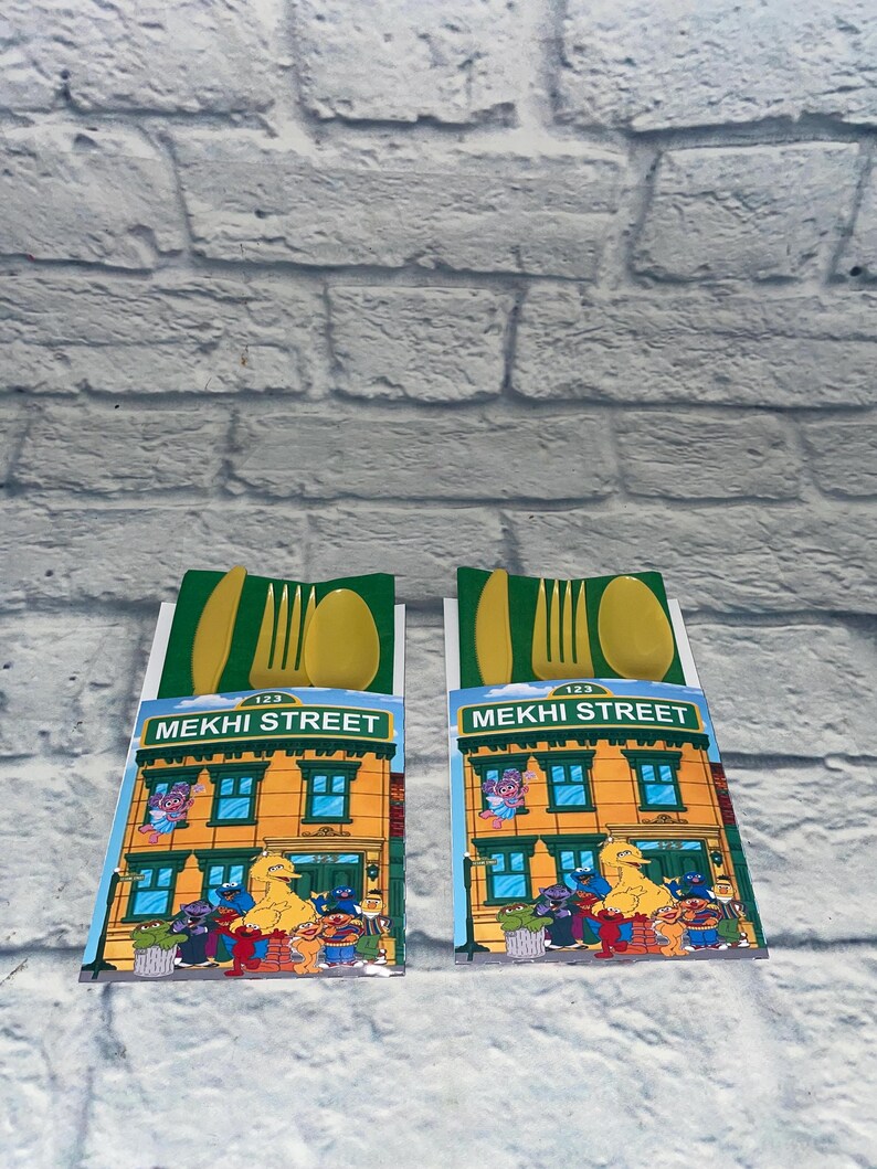 May include: Two sets of yellow plastic cutlery, each packaged with a green and white card featuring a Sesame Street building illustration. The card reads "MEKHI STREET" and includes various Sesame Street characters.