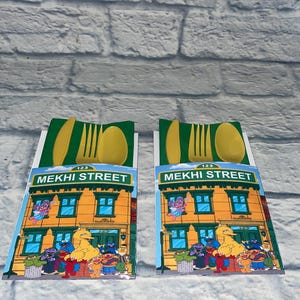 May include: Two yellow plastic cutlery sets. Each set includes a fork, spoon, and knife, presented on a card featuring a colorful illustration of a building with the text "MEKHI STREET" and various cartoon characters.