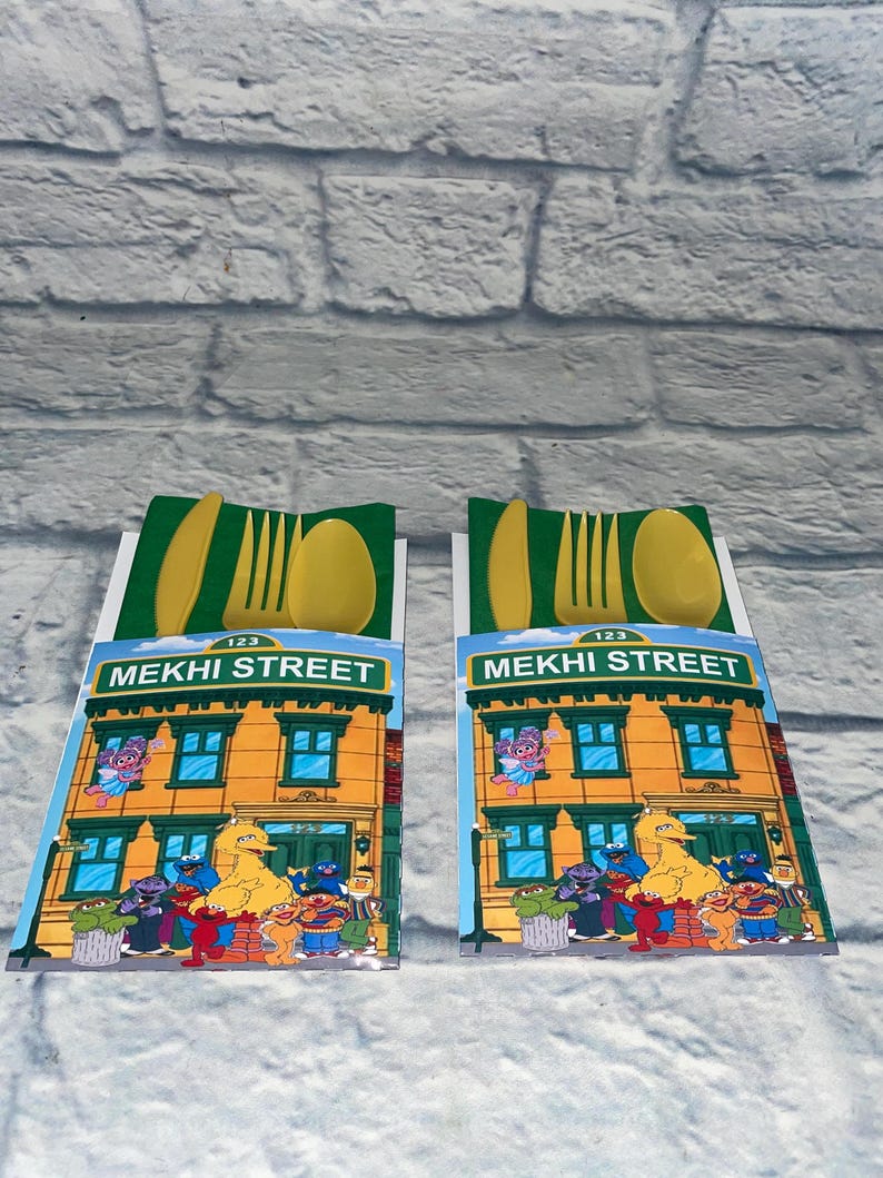 May include: Two yellow plastic cutlery sets. Each set includes a fork, spoon, and knife, presented on a card featuring a colorful illustration of a building with the text "MEKHI STREET" and various cartoon characters.